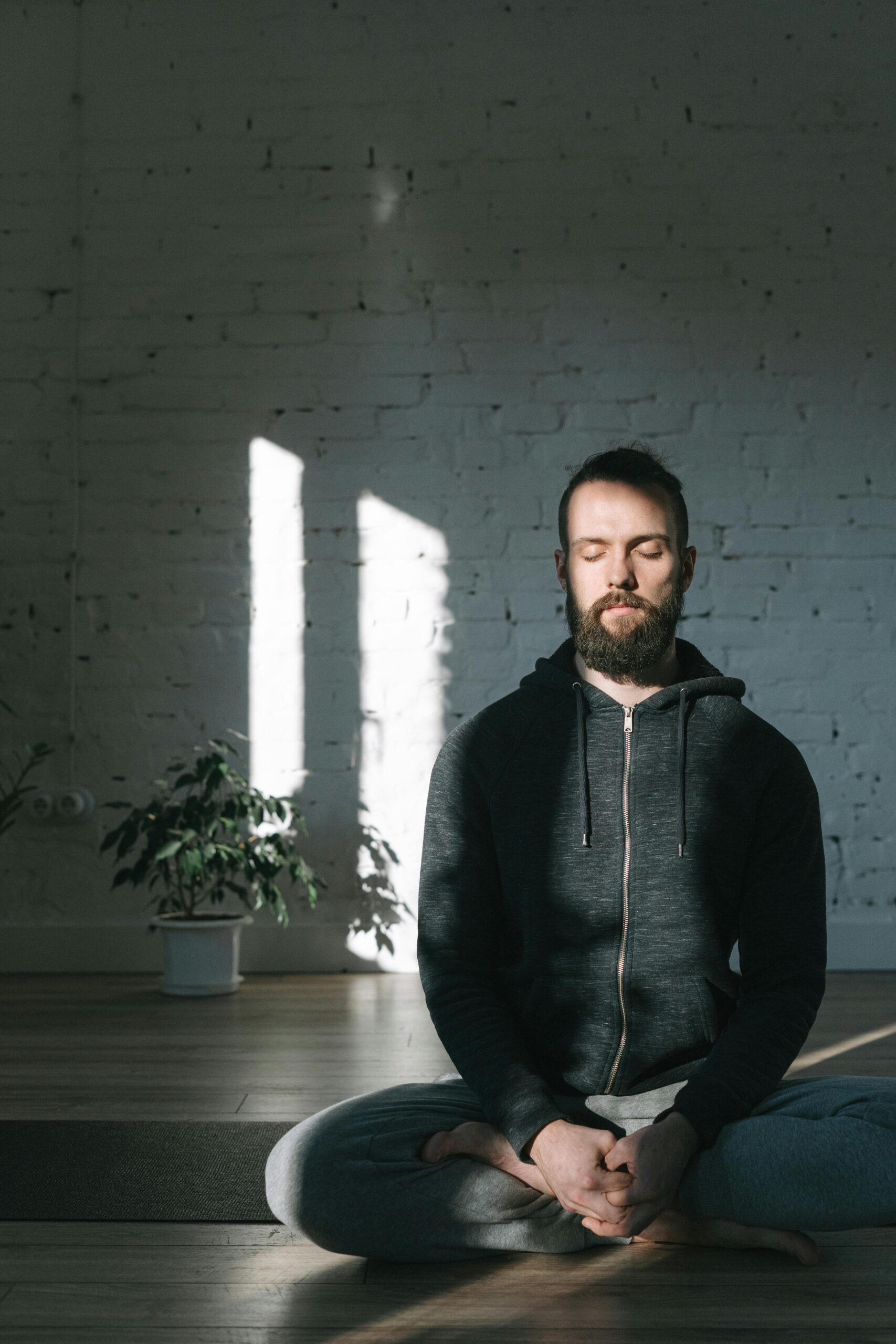 A man in a hoodie meditates in a sunlit yoga studio, embracing tranquility.
